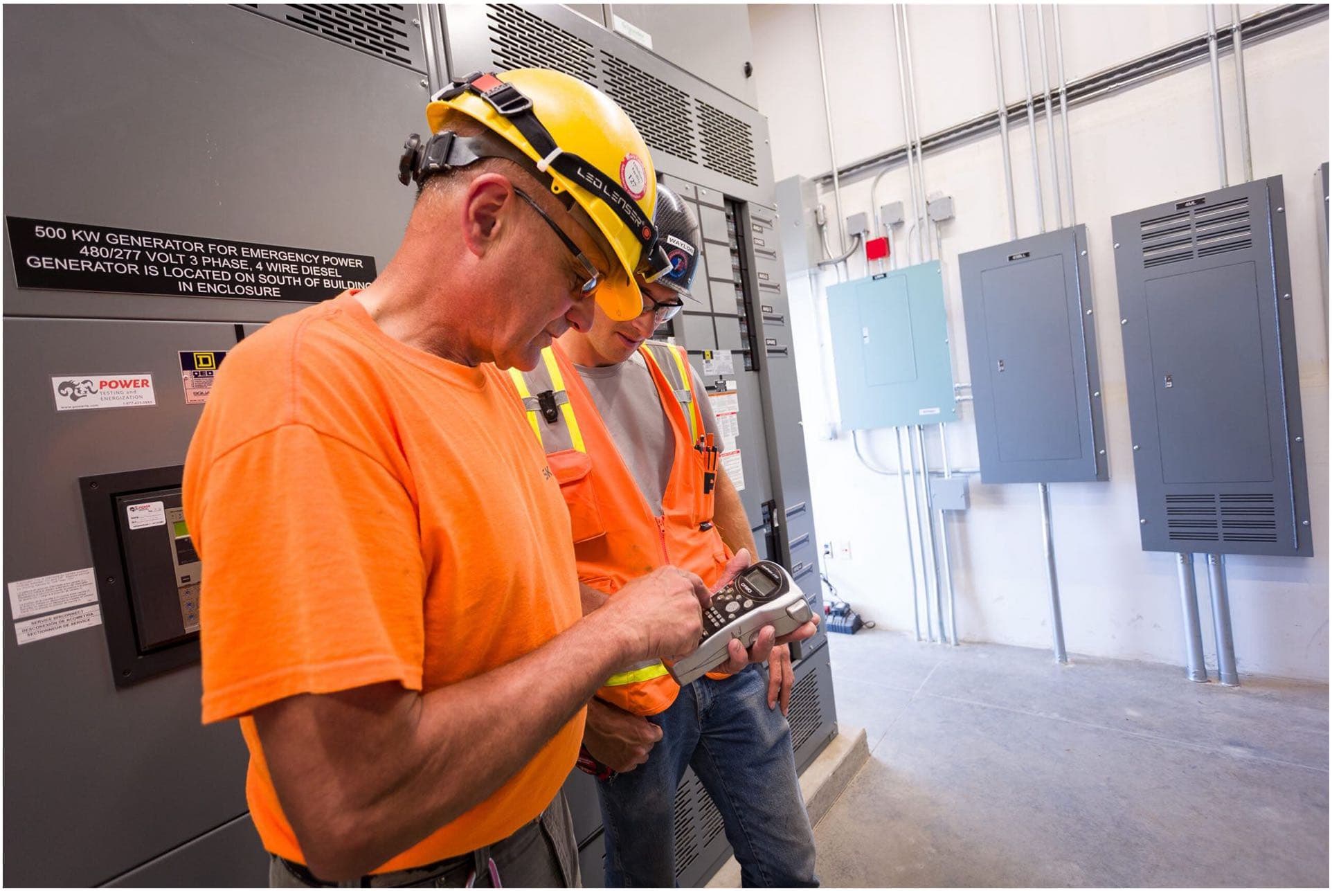 Two workers in safety gear, including hard hats and high-visibility clothing, inspect a device in a control room with electrical panels and a generator. An experienced electrician in Vancouver, WA oversees the detailed inspection to ensure everything operates smoothly.