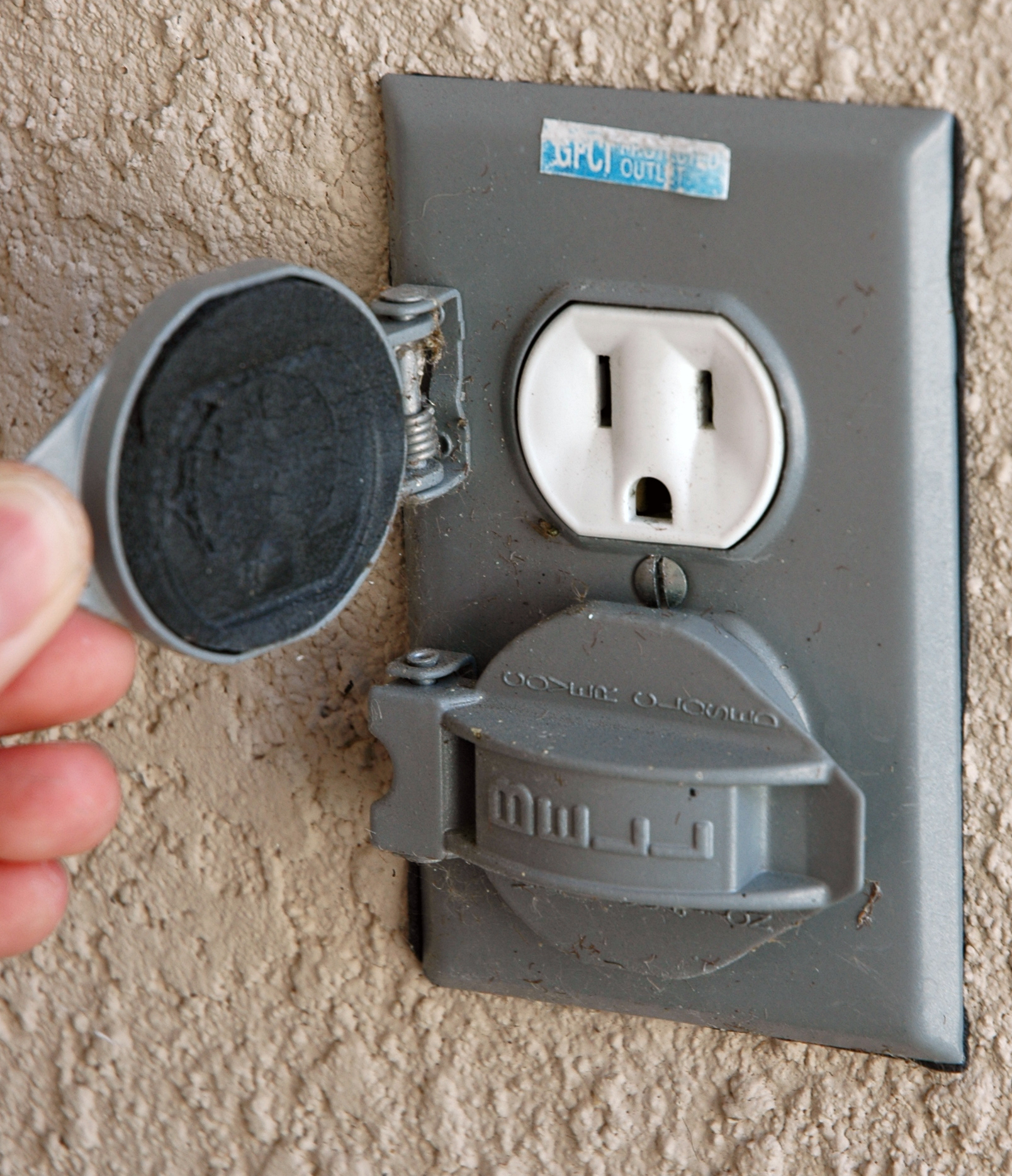 A hand lifts the cover of a gray exterior outlet on a textured wall, exposing a labeled socket—ideal for outdoor outlet repair and electrical troubleshooting in Vancouver, WA.