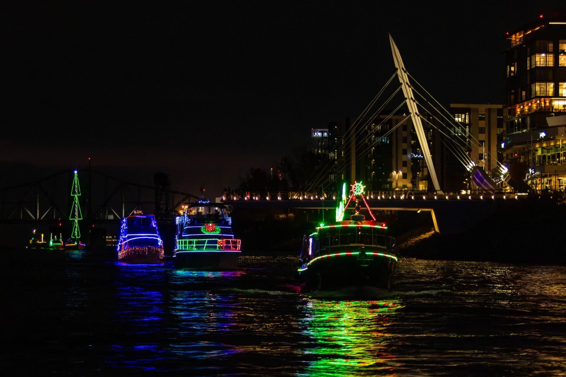 Boats with expertly installed Christmas lights travel a Vancouver, WA river at night; bridge and buildings also feature professional lighting.