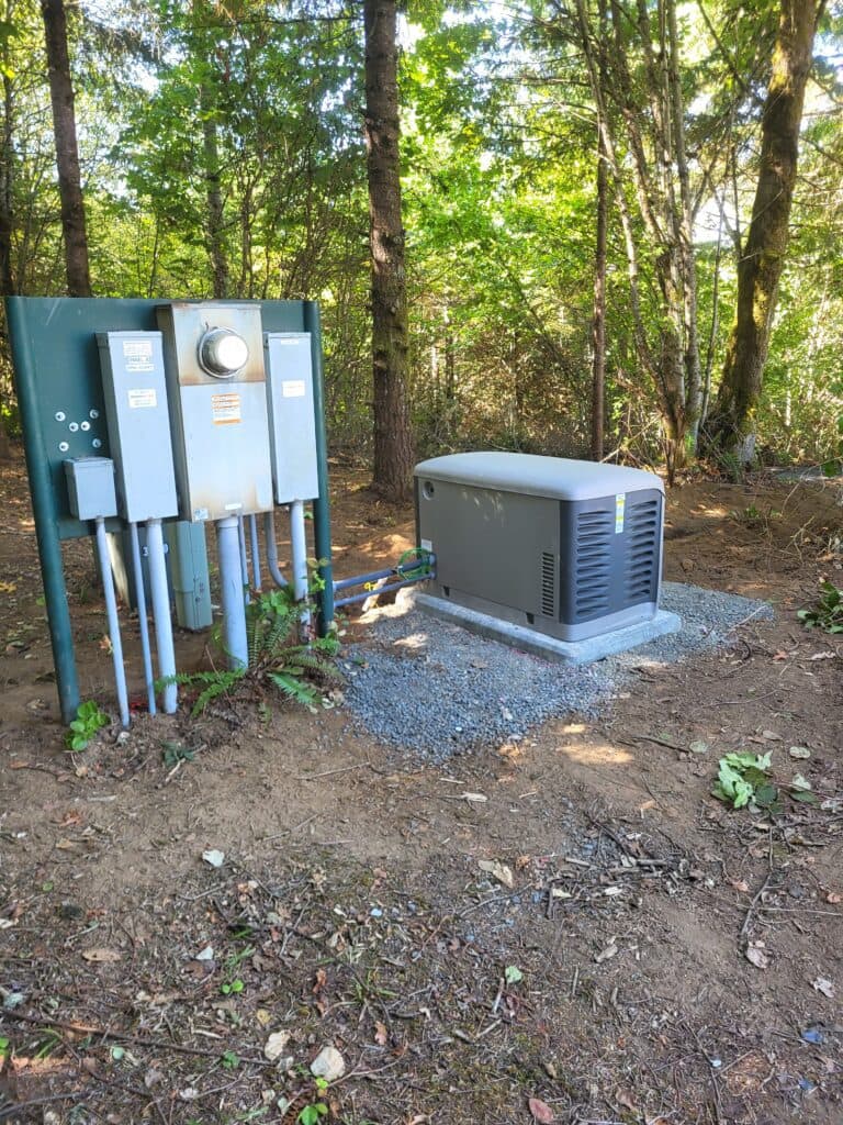 Image shows a metal utility box and a larger generator unit installed on a concrete slab, surrounded by trees and soil with some scattered leaves and branches, showcasing the meticulous work of a Vancouver electrician.