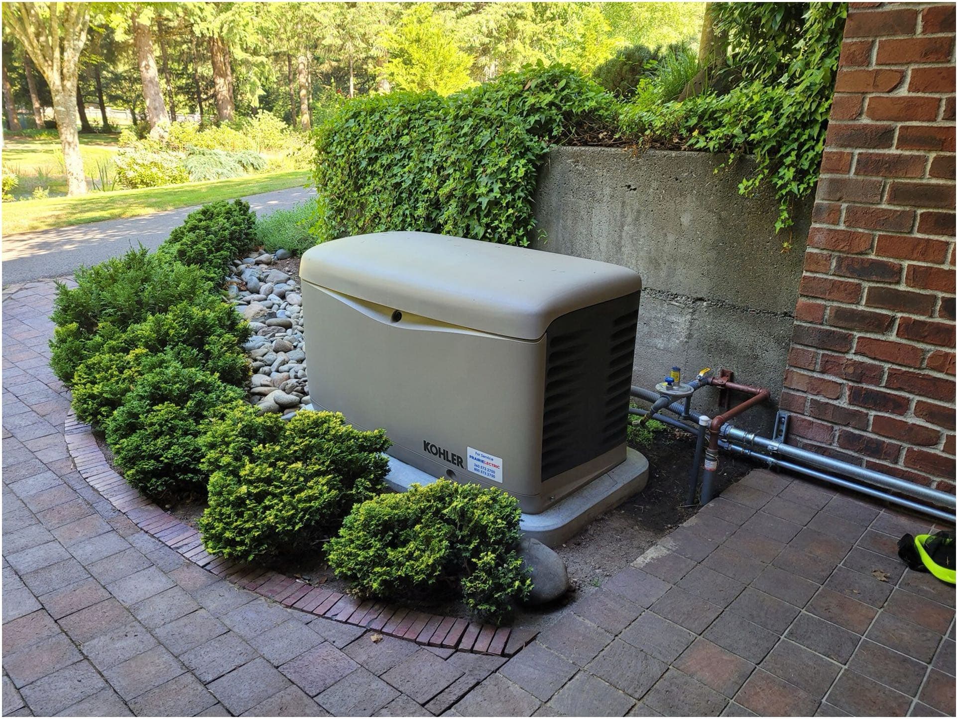 A beige Kohler generator, installed by an electrician in Vancouver WA, sits on a concrete pad next to a brick wall, surrounded by greenery and small shrubs.