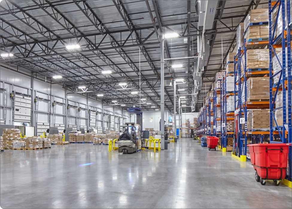 A spacious warehouse interior with large shelves stacked with boxes on the right, and a forklift operating in the middle. Various pallets and containers are scattered throughout the area, while an electrician checks the electrical systems to ensure everything is up to code.