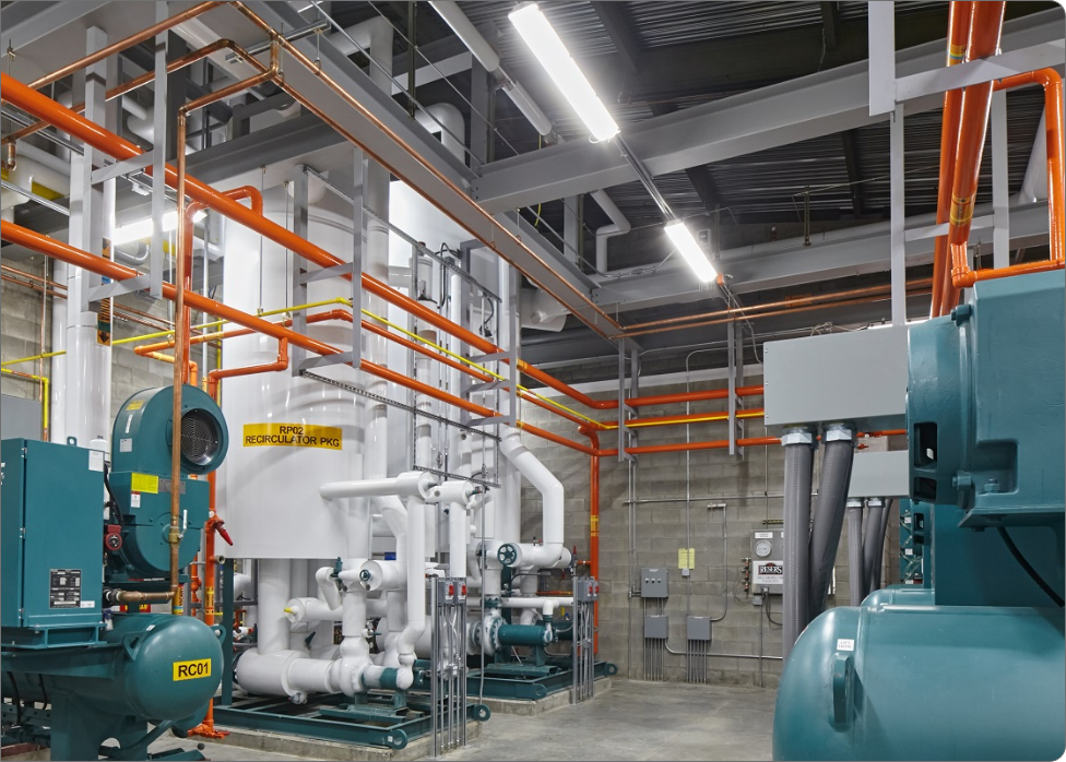 The industrial room, with its complex piping system, large blue machinery, and orange safety railings, showcases the meticulous work of an electrician. Overhead fluorescent lights efficiently illuminate the clean and well-organized space.