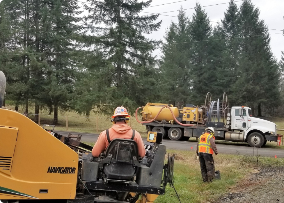 Construction workers are operating heavy machinery near a rural roadside, with a truck and trees in the background. An electrician in Vancouver, WA contributes by ensuring the electrical systems are safely integrated into the project.
