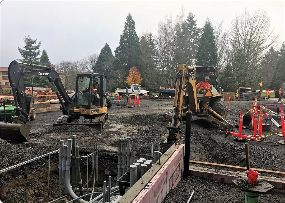 Construction site with two excavators and workers. One excavator is digging while the other prepares to move material beside an electrician inspecting nearby electrical setups. Various construction materials and safety cones are visible around the site.
