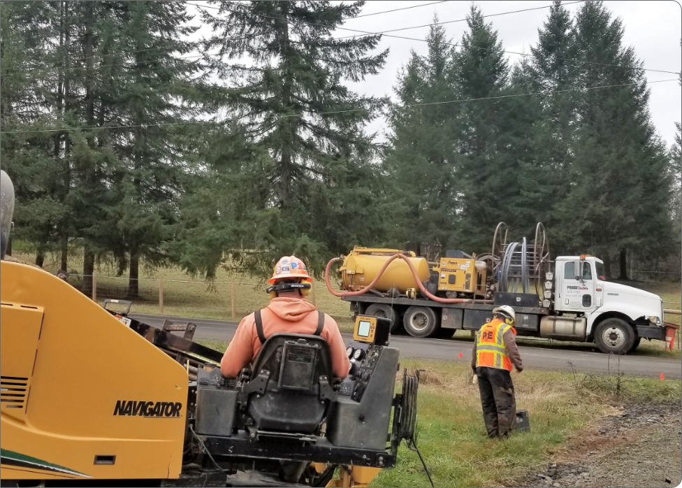 Construction workers operate heavy machinery on a roadside near a forested area, with a yellow piece of equipment and a white truck in the background. Nearby, a Vancouver electrician is seen inspecting the electrical systems to ensure safety for the ongoing work.