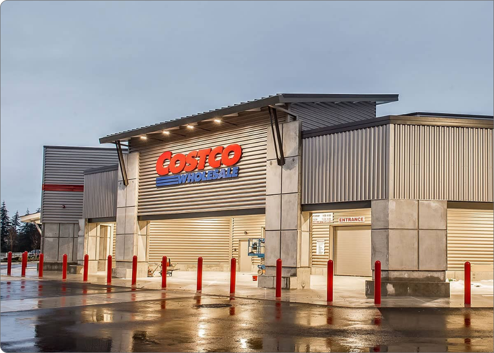 The exterior of a Costco Wholesale warehouse, featuring the main entrance with illuminated signage and red bollards in front, looks vibrant even after recent rain. If you're near Vancouver, WA and need expert electrical services, finding a skilled electrician here is just as seamless.