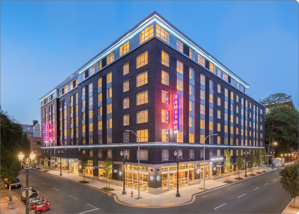 A large six-story brick hotel with illuminated signs on the corner of two streets at dusk, featuring lit windows maintained by a Vancouver electrician and a well-lit ground floor entrance.