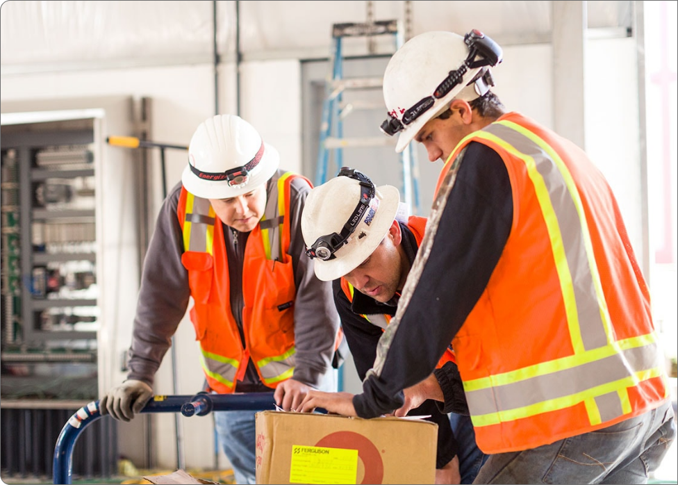 Three construction workers wearing white hard hats and orange safety vests inspect a cardboard box. One worker, an electrician, points at the box while the others look on attentively.