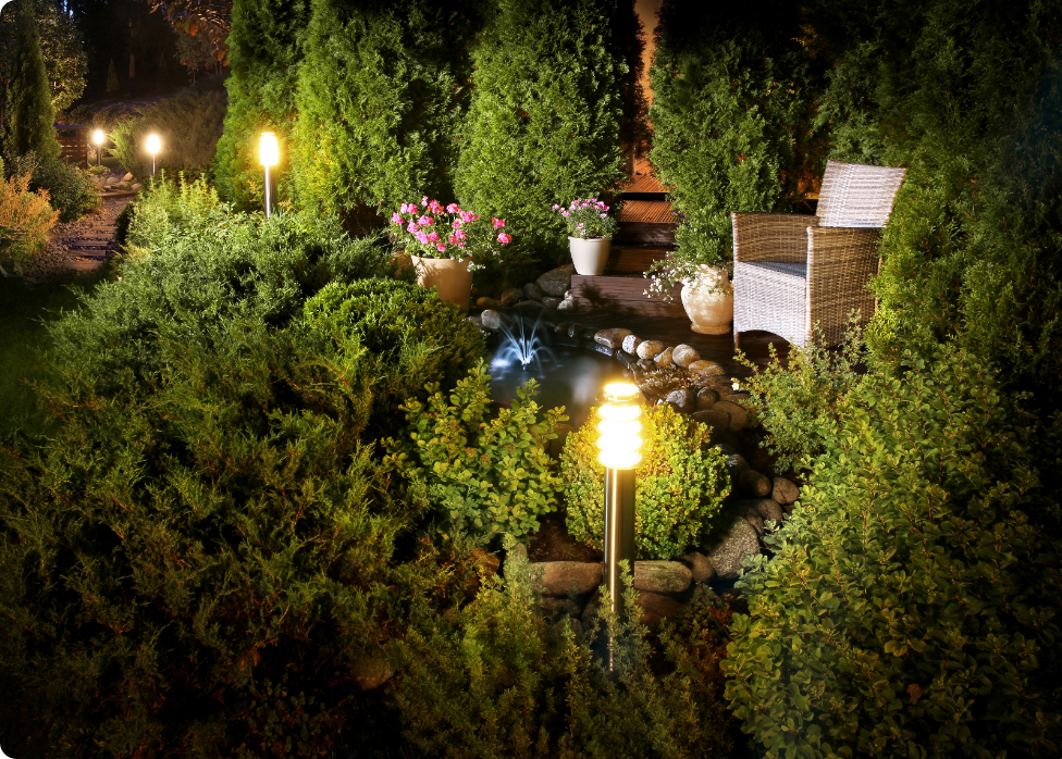 A well-lit garden scene at night featuring a wicker chair, potted flowers, shrubs, and a small pond with surrounding greenery—beautifully illuminated thanks to an expert electrician in Vancouver, WA.