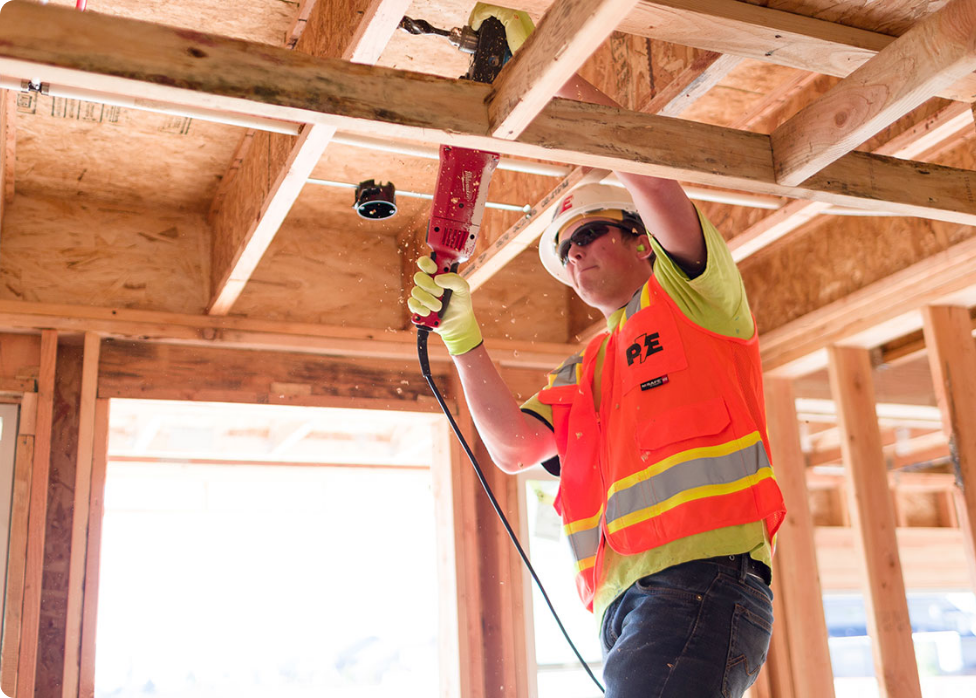 A construction worker in a safety vest and helmet uses a power tool to drill into a wooden frame, while an electrician in Vancouver WA wires the electrical system nearby, ensuring the building under construction is both solid and functional.