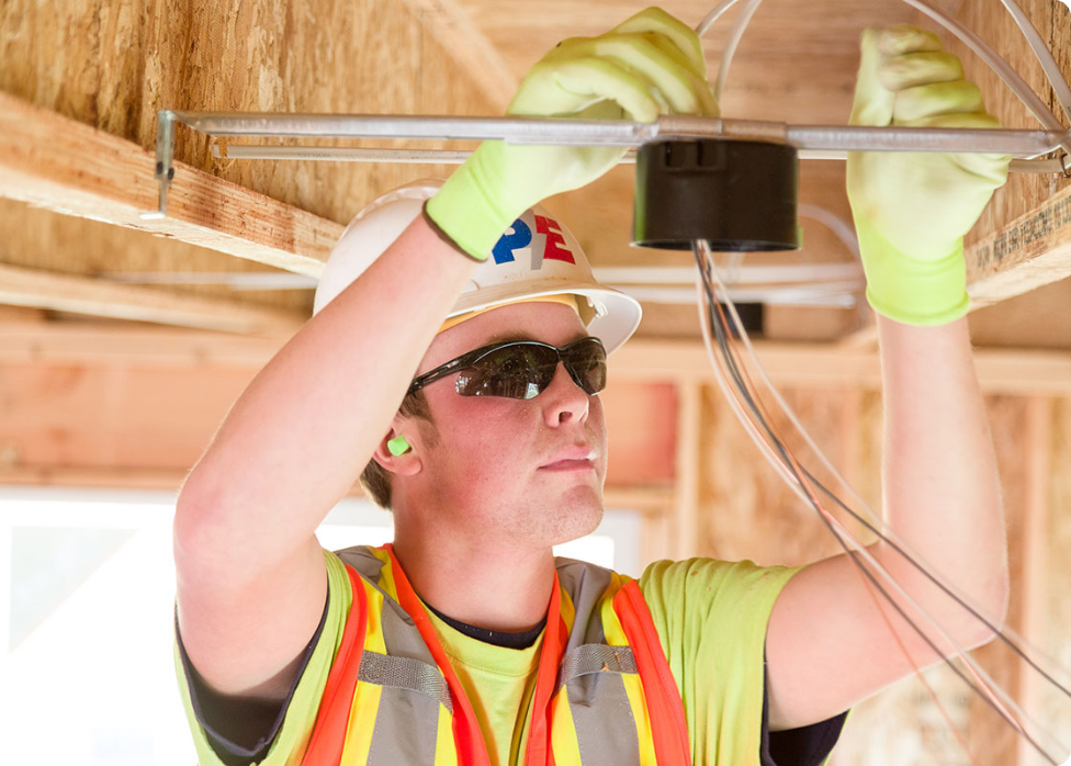 An electrician in Vancouver, WA, wearing a hard hat, safety glasses, and gloves installs electrical wiring in the ceiling of a wooden structure.
