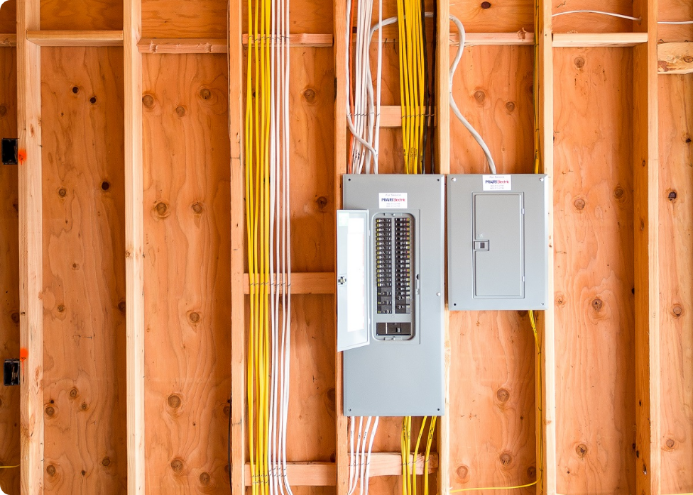 Two electrical panels and several yellow and white cables are mounted on a wooden wall. One panel door is open, revealing the circuit breakers inside. If you need assistance, consider contacting an experienced Vancouver electrician to ensure everything is up to code.