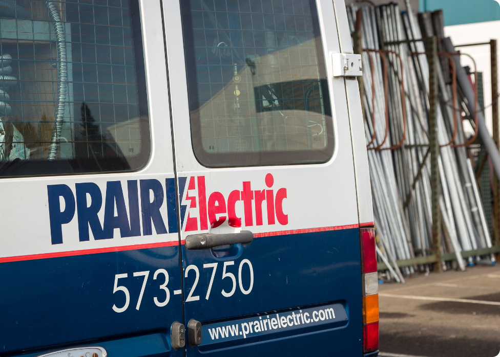 A van with "Prairie Electric" signage and contact number, belonging to an experienced electrician in Vancouver, WA, parked next to a stack of metal pipes.