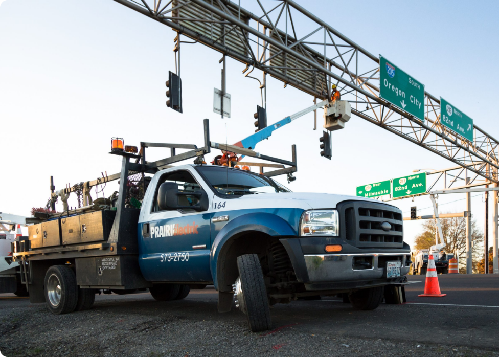 A work truck with "Prairie Electric" on the door is parked under traffic lights and highway signs as an electrician in Vancouver, WA performs maintenance using a cherry picker.