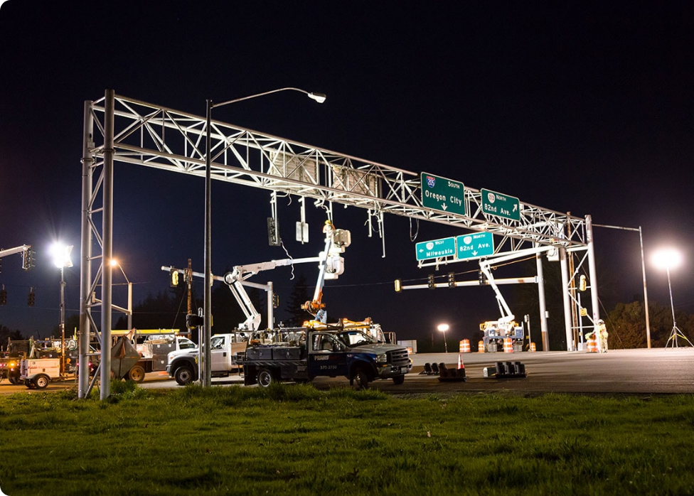 Workers in elevated buckets, including a skilled Vancouver electrician, perform maintenance on overhead traffic signs on a roadway at night, with vehicles and equipment present.
