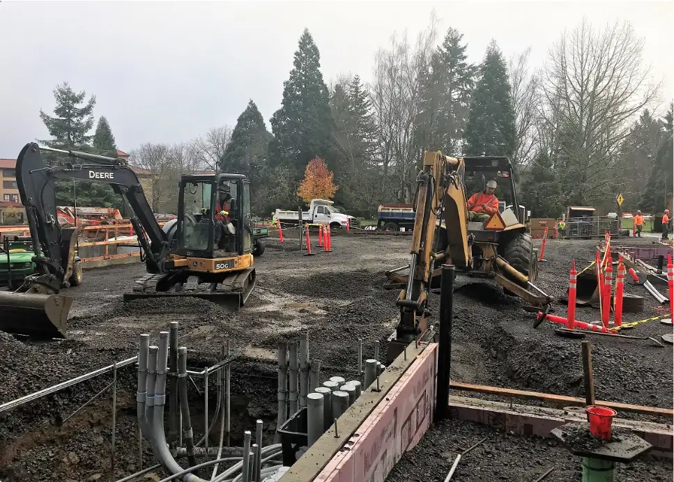 Construction site with two excavators, workers in safety gear, and utility pipes visible. An electrician in Vancouver, WA is inspecting the electrical setup. Trees and construction vehicles are in the background.