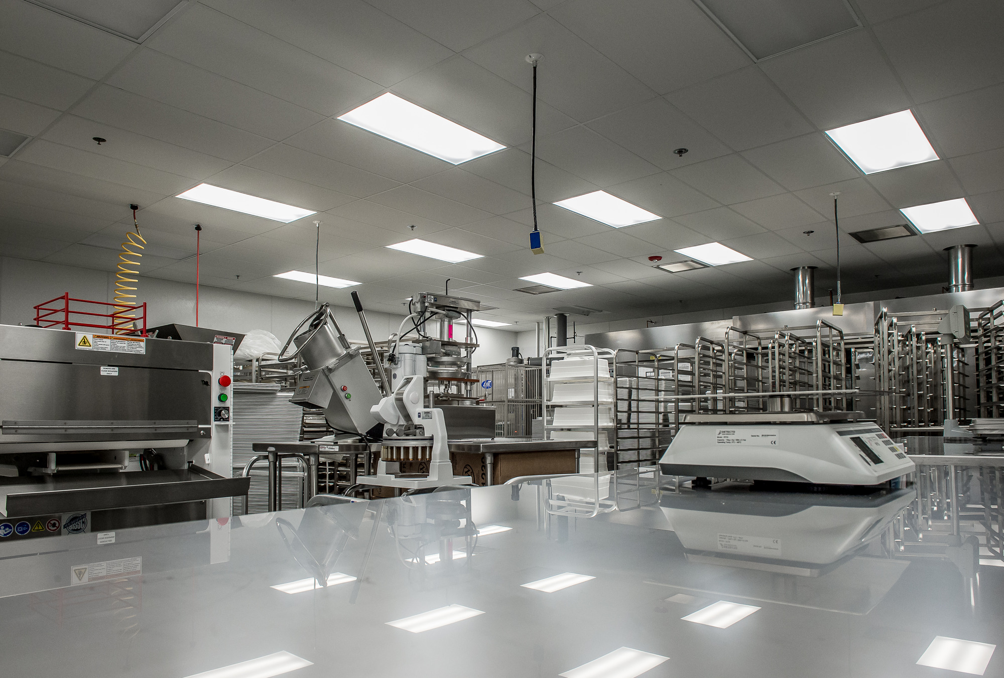 An industrial kitchen in Bellingham, WA, features stainless steel equipment, racks, and preparation tables. Overhead lights brighten the space for efficient operations. For electrical maintenance and installation services, consider hiring a professional electrician to ensure optimal lighting and power supply.