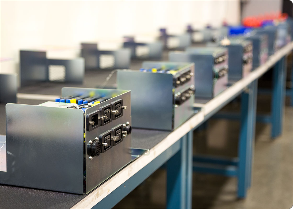 A row of partially assembled electronic devices in metal cases is lined up on a long workbench in a factory, meticulously crafted by skilled Vancouver electricians.