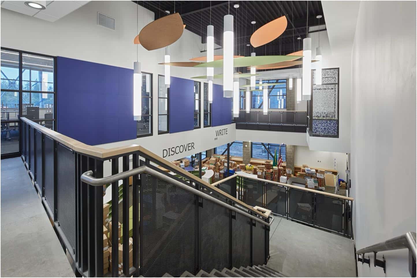 Interior view of a modern, multi-level library with stairs leading down to a main area. Bookshelves, study areas, and large windows are visible, along with words "DISCOVER" and "WRITE" on a wall.