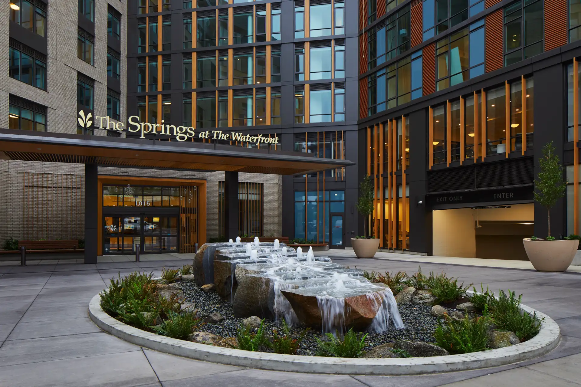 Fountain in front of a modern building entrance with a sign reading "The Springs at The Waterfront." The setting includes large windows, planters, and directional signs for exit and parking.