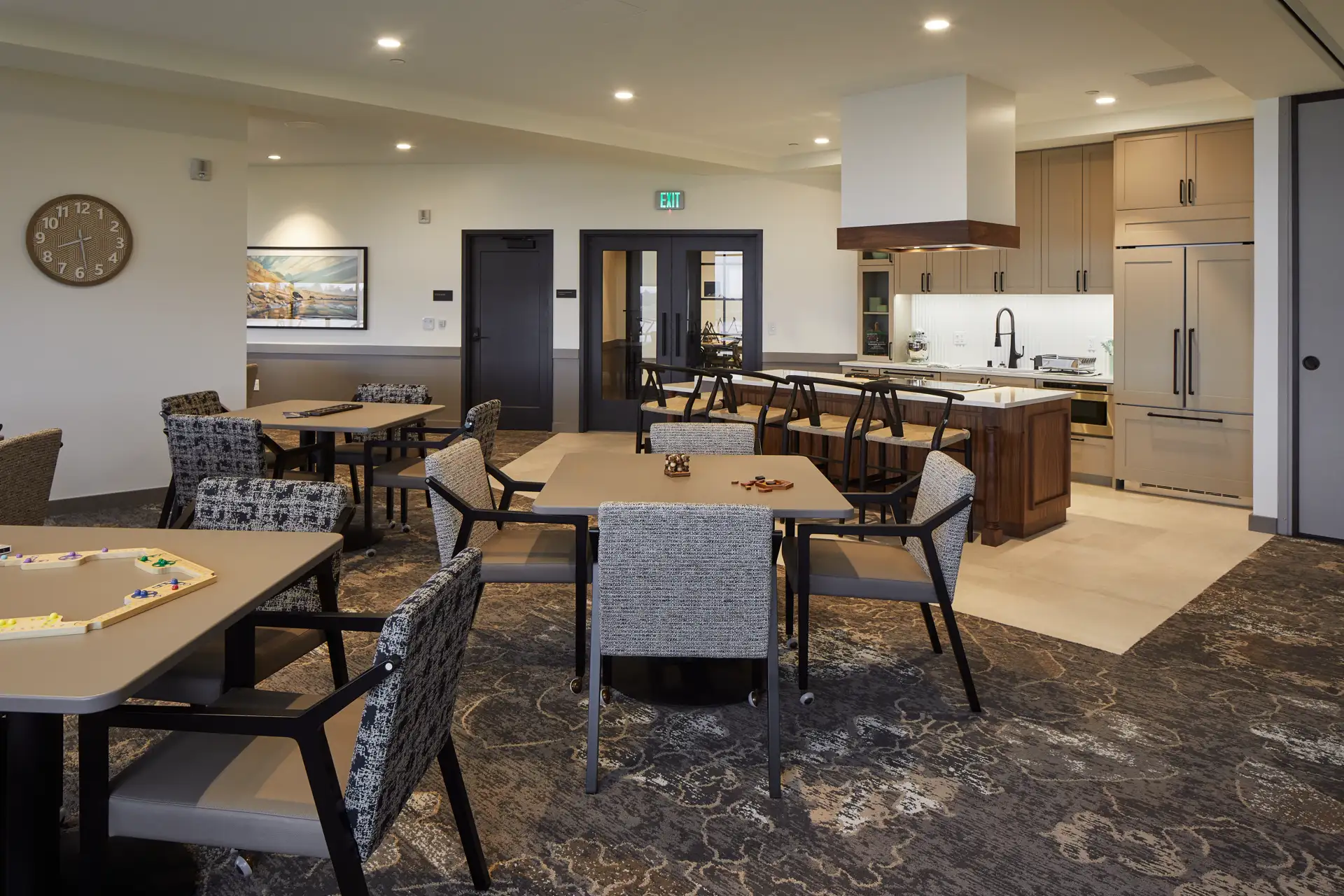 A modern dining area with several tables and chairs, a kitchen island with stools in the background, and decorative lighting in an open, well-lit space.