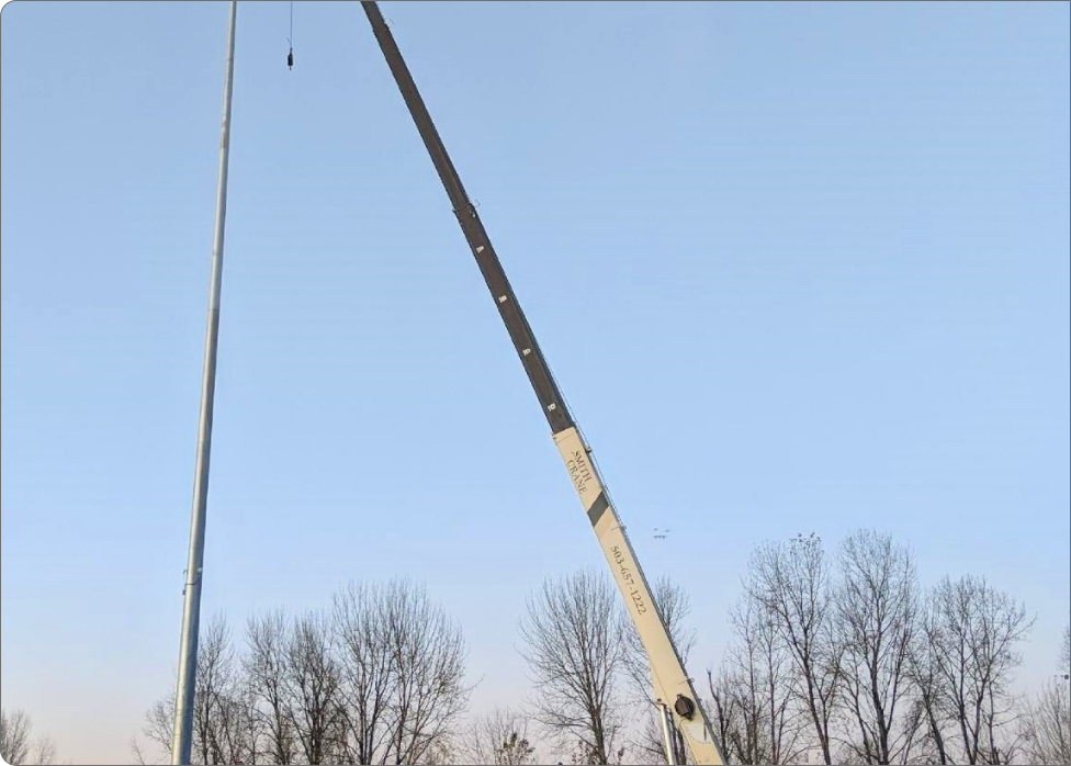 A crane is lifting a tall metal pole in an outdoor area with trees in the background. The sky is clear and blue, making it a perfect day for the work of an electrician in Vancouver WA.