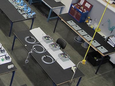 A workspace with several workstations equipped with electrical devices, wires, and components. Some tables have open toolkits and scattered documents. A yellow cord hangs from the ceiling, reminiscent of an electrician's setup in Vancouver, WA.