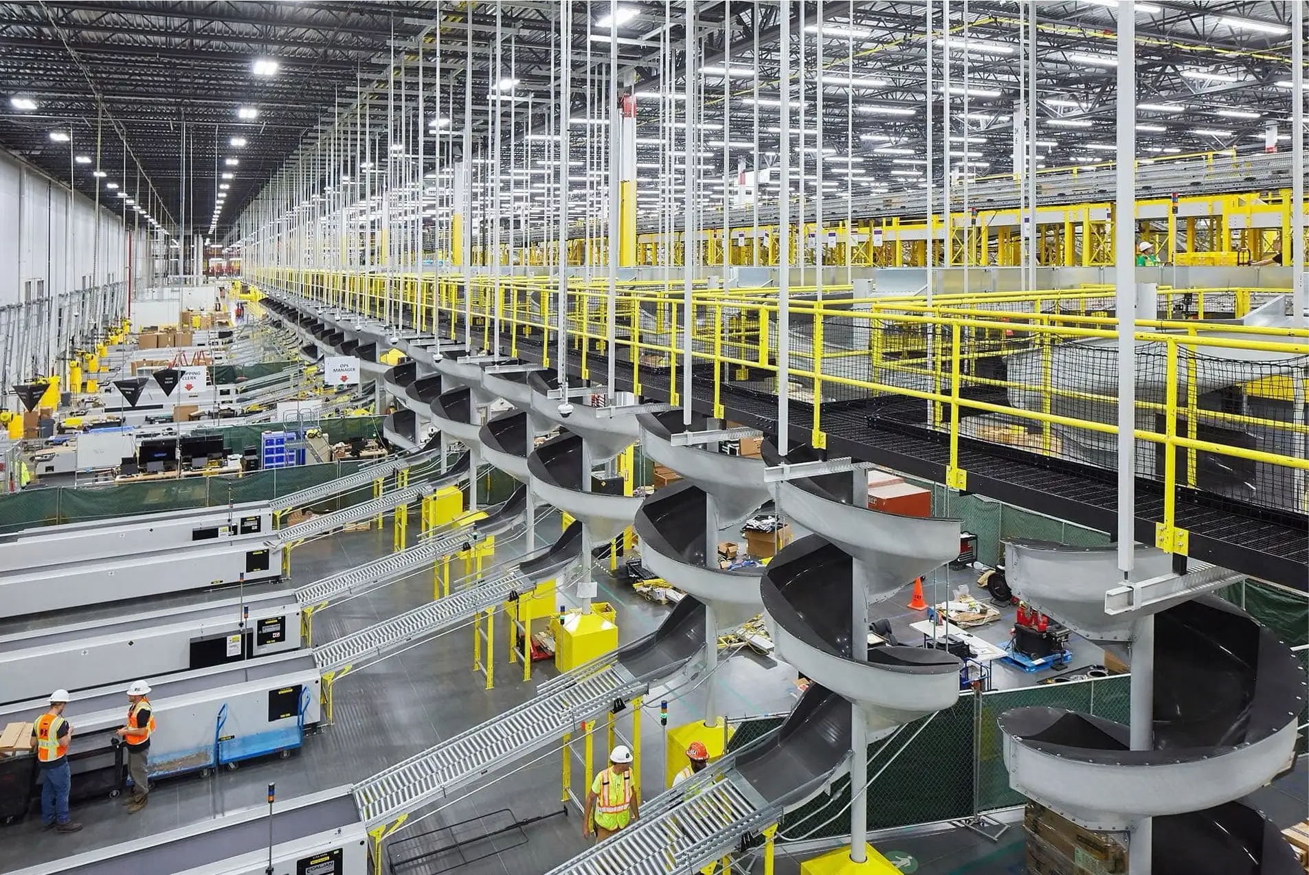 A large warehouse with multiple conveyor belts and sorting chutes. Several workers in safety vests and helmets are present, alongside an electrician ensuring all electrical systems run smoothly. The space is brightly lit with overhead lighting and extensive infrastructure.