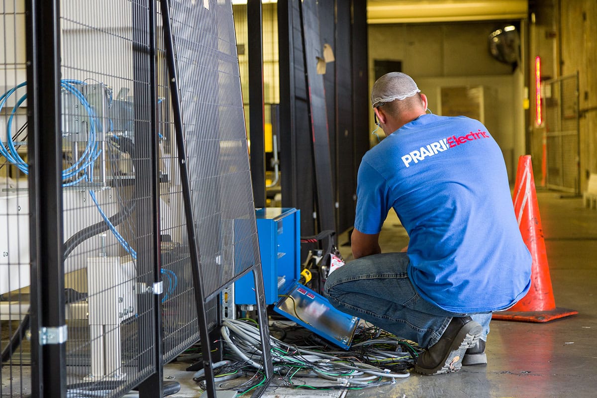 A person in a blue "PRAIRIElectric" shirt crouches while working near electrical cables and equipment in an industrial setting. An orange traffic cone is visible nearby.