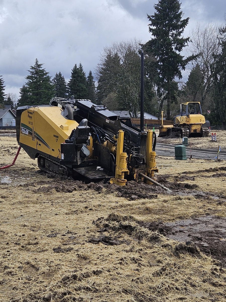 A yellow horizontal directional drilling machine operates on a muddy construction site in Vancouver, WA, surrounded by trees and residential buildings. Nearby, a bulldozer is parked under a cloudy sky.