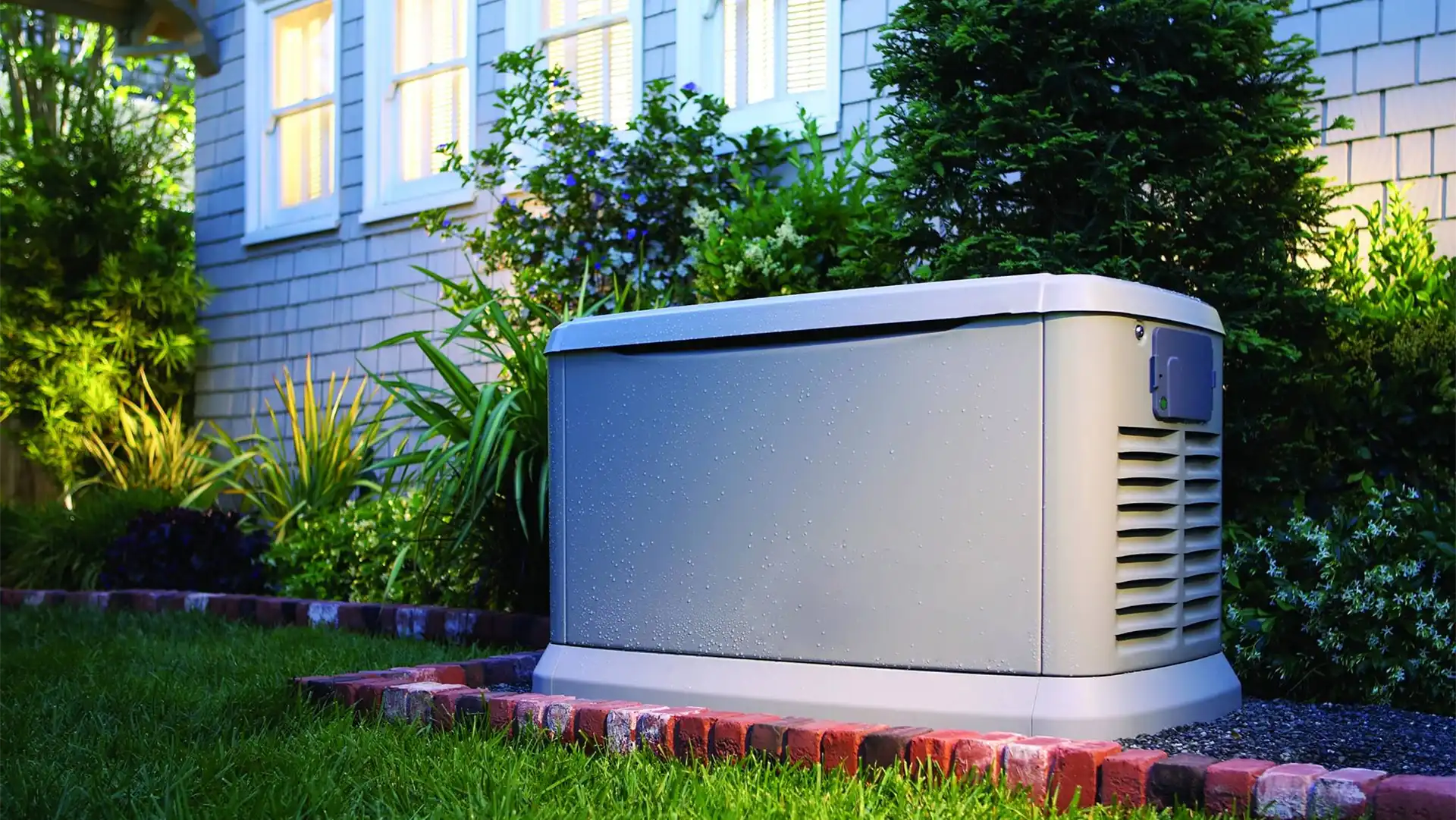 A gray standby generator installed on a brick platform outside a house with lit windows and surrounding vegetation, expertly set up by a skilled Vancouver electrician.
