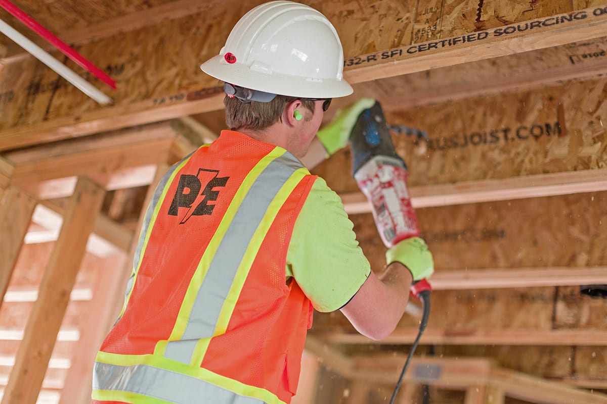 A construction worker in a hard hat and high-visibility vest operates a power tool while working on wooden framing at a site, alongside a Vancouver electrician handling the electrical wiring.