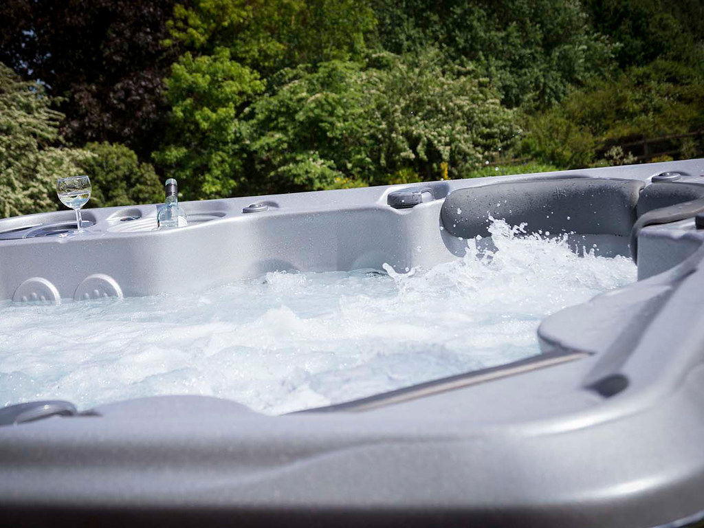 A bubbling hot tub outdoors with greenery in the background and a glass of white wine placed on the edge.
