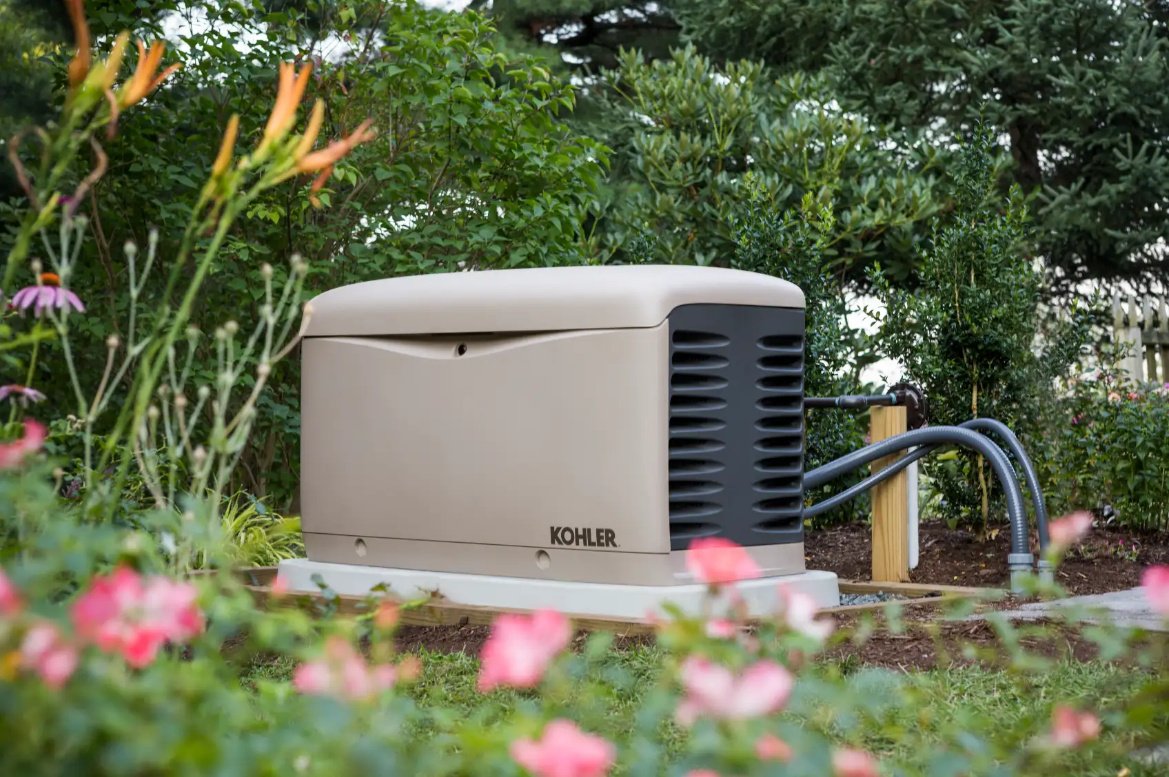An outdoor backup generator labeled "Kohler" installed on a concrete pad, surrounded by bushes and flowers, might be the handy work of a skilled electrician in Vancouver, WA.