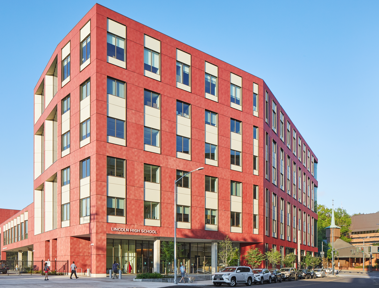 A modern, red and beige multi-story building with large windows labeled "Lincoln High School," featuring people, bicycles, and an electrician working on a nearby electrical panel in front.