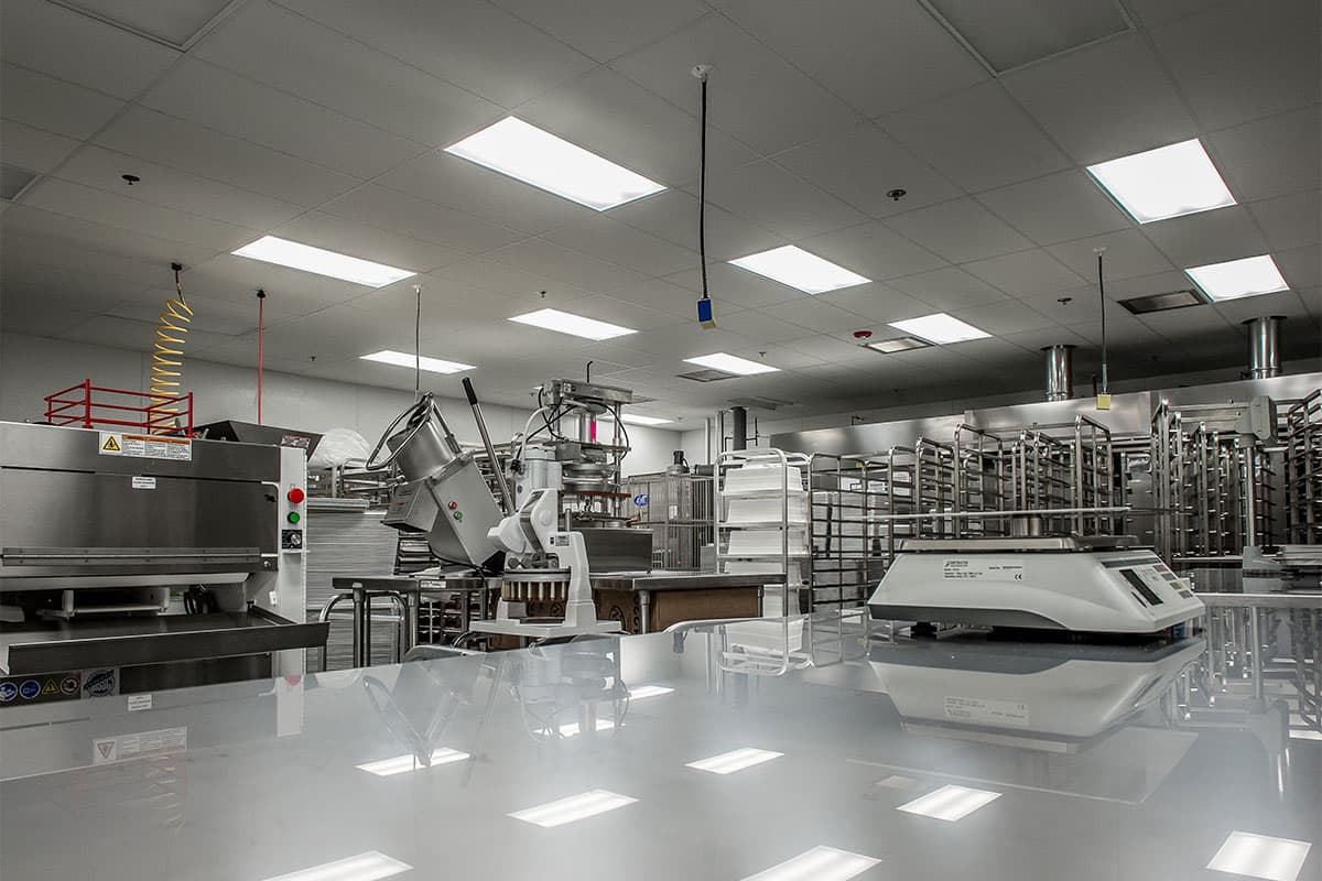A large, well-lit commercial kitchen with stainless steel equipment, including ovens, racks, and workstations. Bright overhead lights installed by an electrician reflect on the shiny surfaces.
