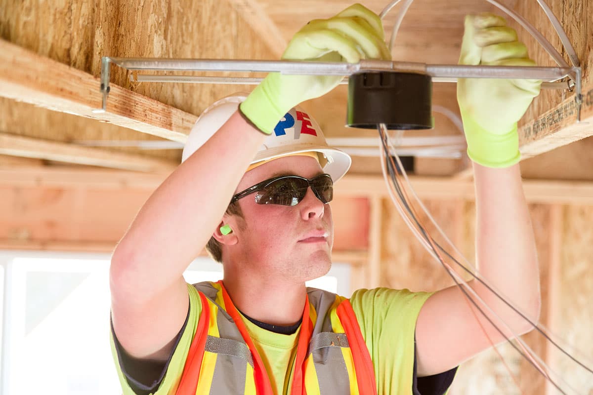 A construction worker wearing a hard hat, safety vest, gloves, and sunglasses installs electrical wiring in a wooden frame structure.