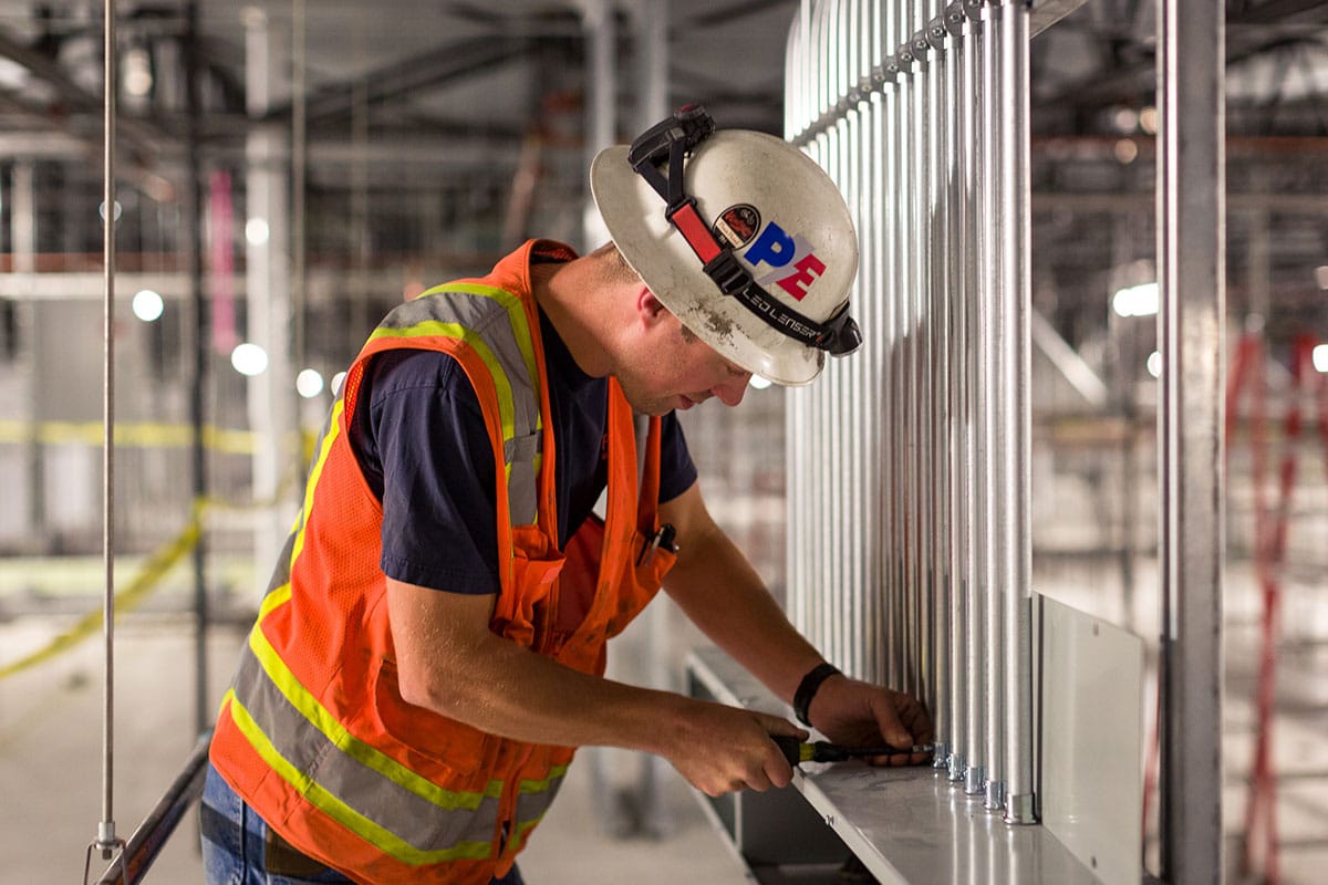 A construction worker in a hard hat and safety vest installs piping alongside an electrician in an industrial setting.