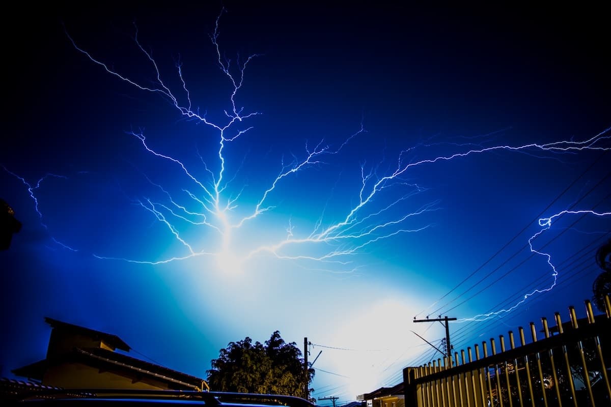 A night sky illuminated by multiple lightning bolts, with trees and rooftops silhouetted in the foreground, likely causing power surges, and reminiscent of the power harnessed by an electrician in Vancouver, WA.
